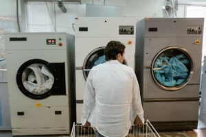 pexels-photo-8774371 - Spinz - Spinz Man in a white shirt seen from behind stands in front of two large industrial washing machines with clothes inside the open door on the right in a laundromat setting | Spinz | Spinz