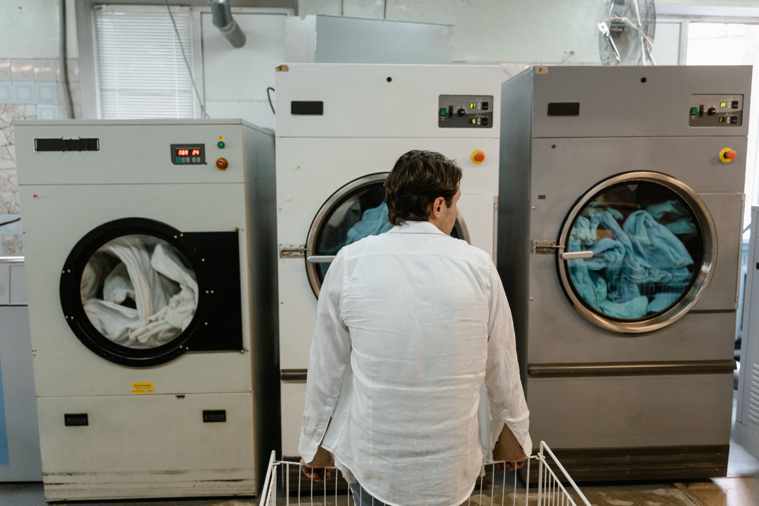 Man in a white shirt seen from behind stands in front of two large industrial washing machines with clothes inside the open door on the right in a laundromat setting